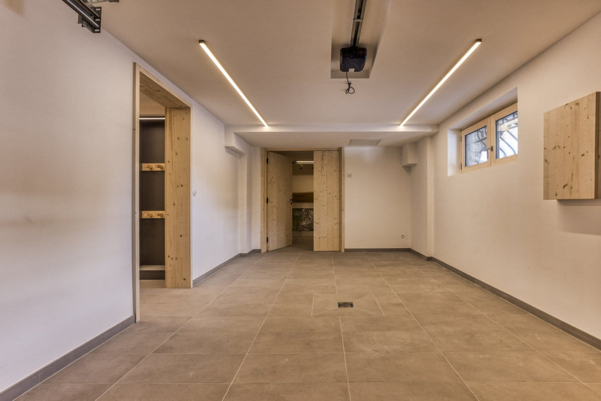 An empty modern garage or storage room with light gray tiled floors, white walls, recessed LED strip lighting, and unfinished light wood closet doors and wall panels.