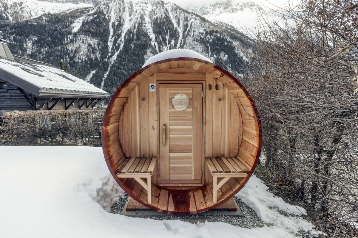 A round wooden sauna cabin with a slatted door and built-in benches, set outdoors in deep snow with mountains and bare trees in the background.