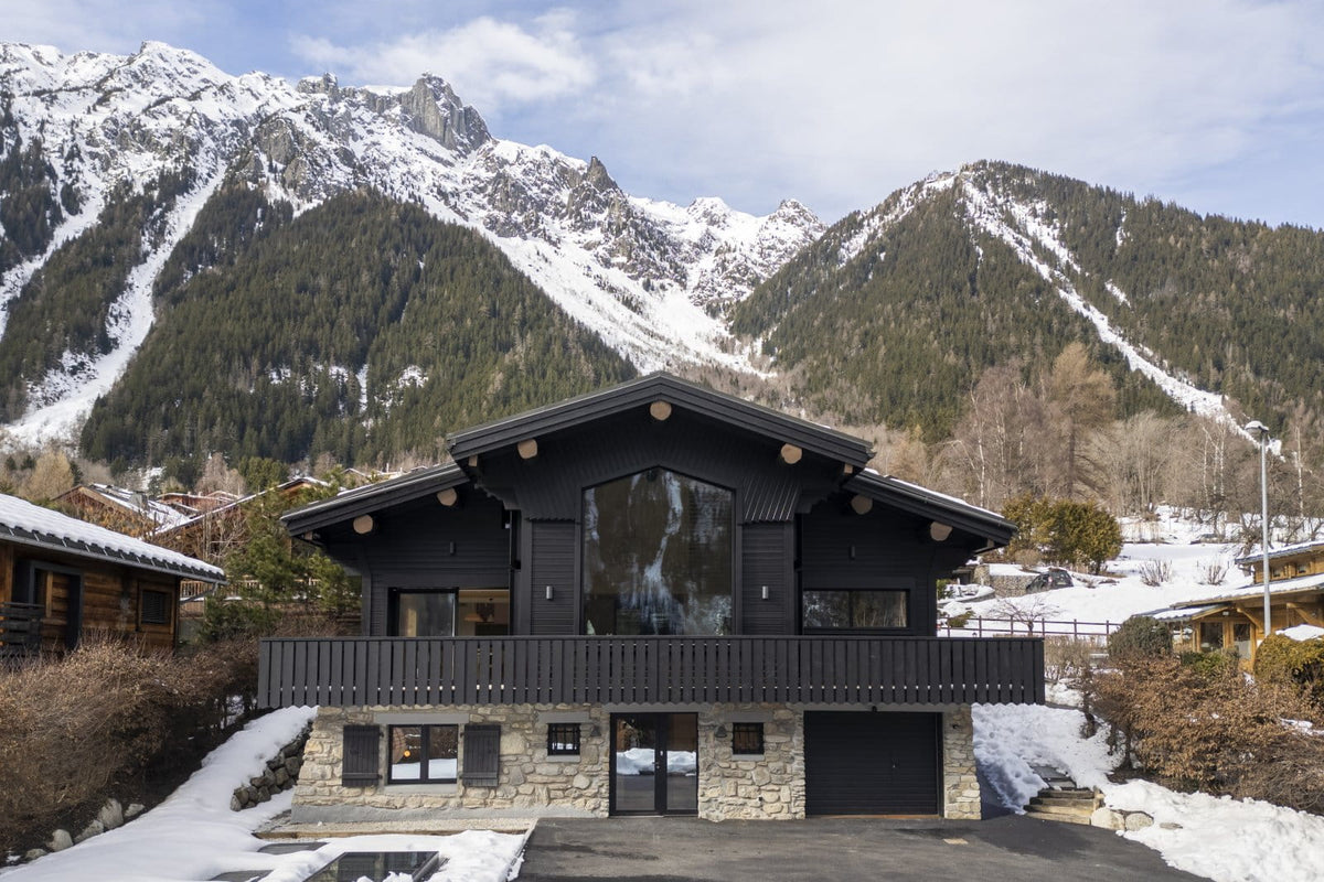 A modern black wooden chalet with a stone base, set against a dramatic backdrop of snow-covered mountains and pine forests.