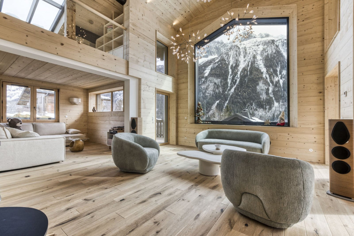 A modern high-ceilinged wooden chalet living room with light wood plank floors and walls, featuring a large black-framed window showing a snowy mountain scene. In the foreground, two sculptural light gray upholstered armchairs surround a low white coffee table.