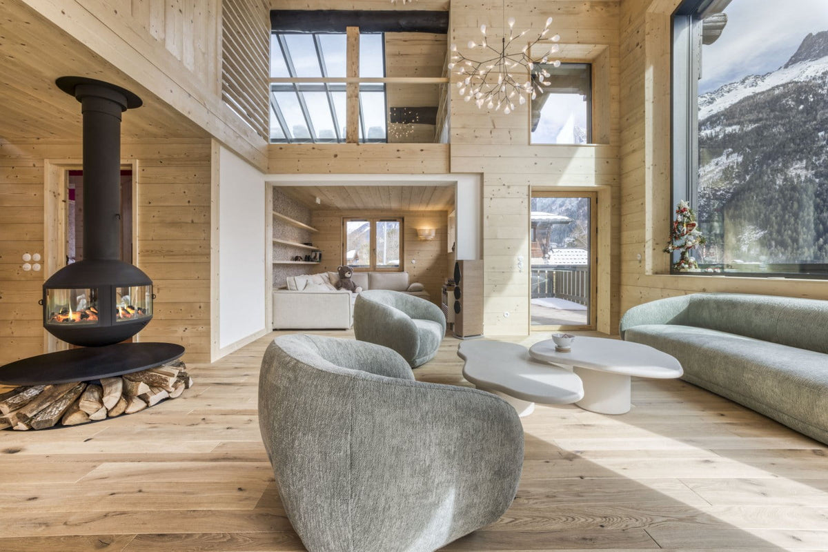 A modern mountain chalet living room with light wood walls and floors, featuring a black freestanding wood-burning stove, two gray upholstered armchairs, a curved sofa, and large windows overlooking snowy mountains.
