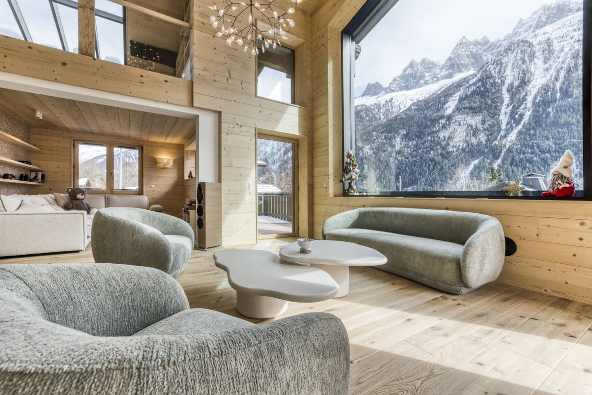 A modern high-ceilinged chalet living room with light wood walls and floor, featuring plush gray upholstered armchairs, a matching curved sofa, white sculptural coffee tables, and a massive window overlooking snowy mountains.