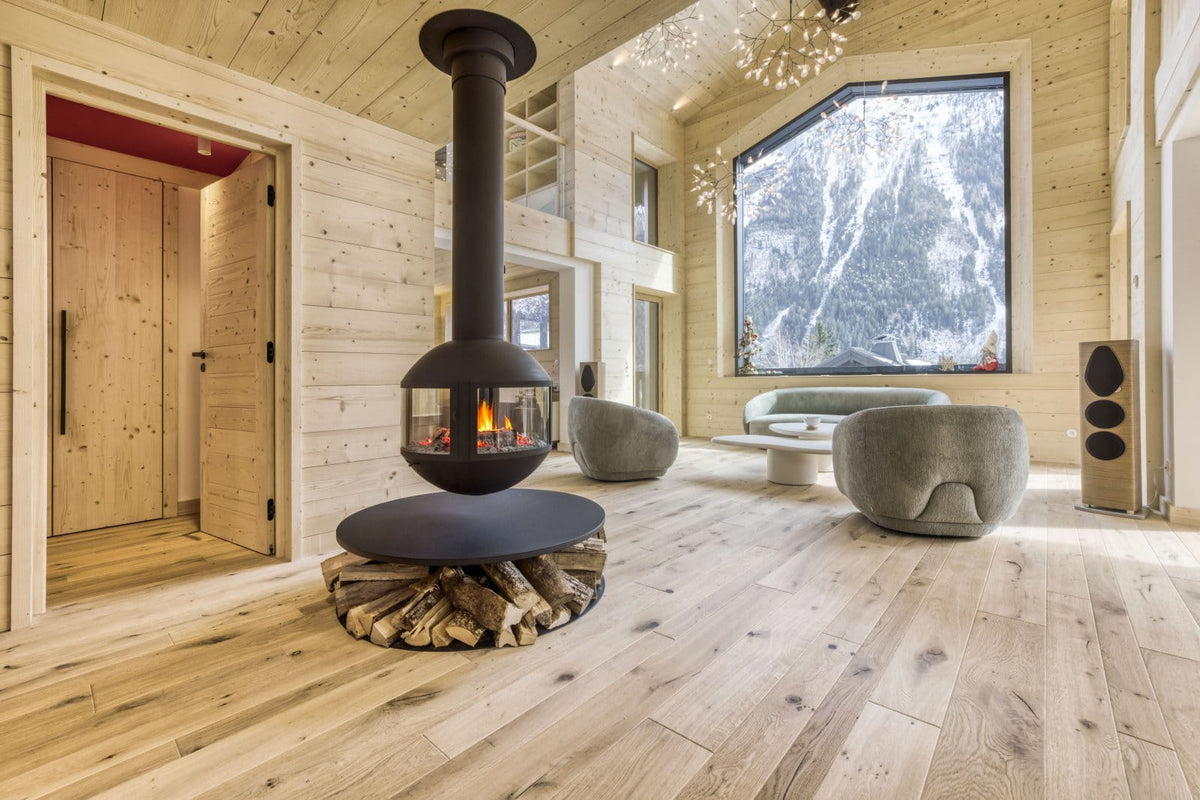 A modern black wood-burning fireplace with a log base stands in a light wood-paneled mountain chalet living room with a large window showing a snowy outdoor view.