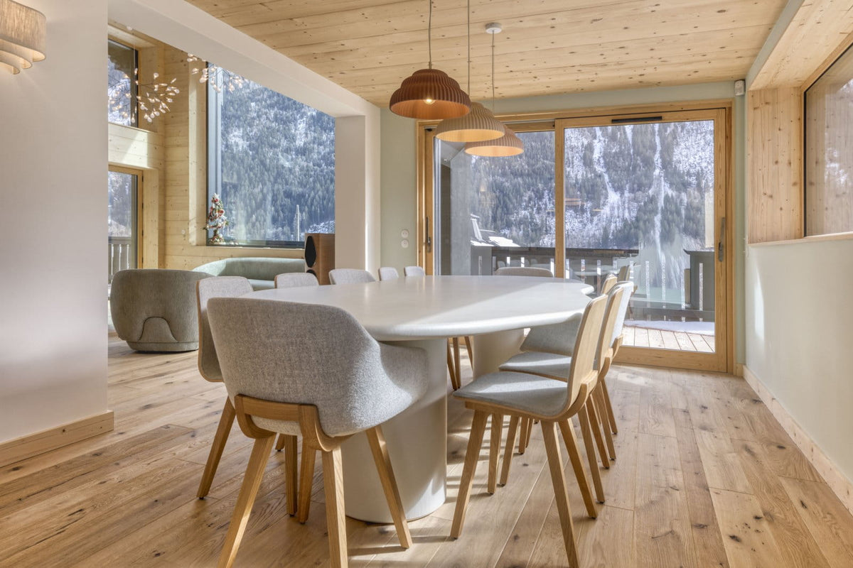 A modern dining room with a round white table and six upholstered dining chairs with light wood legs, set in a bright room with light wood walls and ceiling, large windows, and a view of a snowy outdoor landscape.