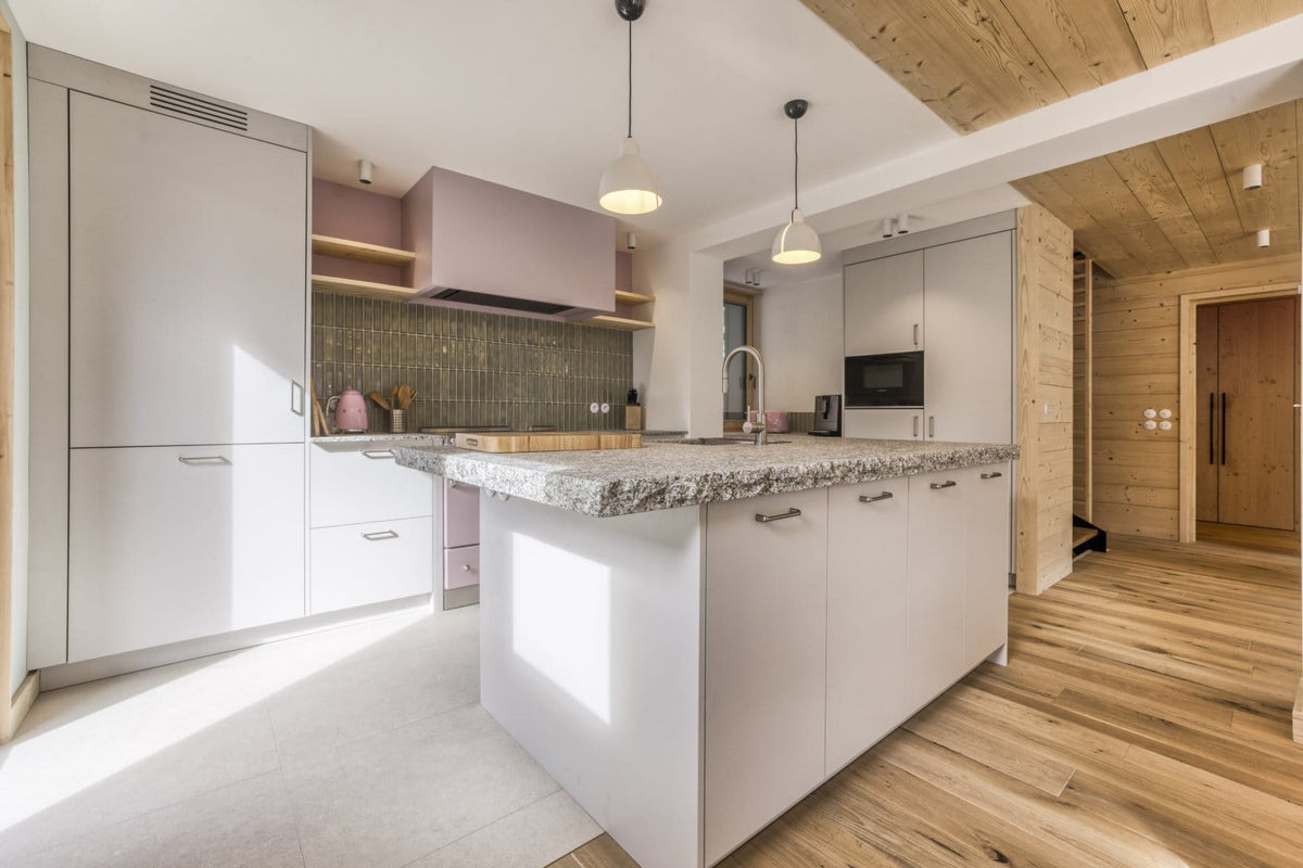 A modern kitchen interior with white cabinetry, a central island with a speckled stone countertop, wood flooring, and light wood ceiling panels.