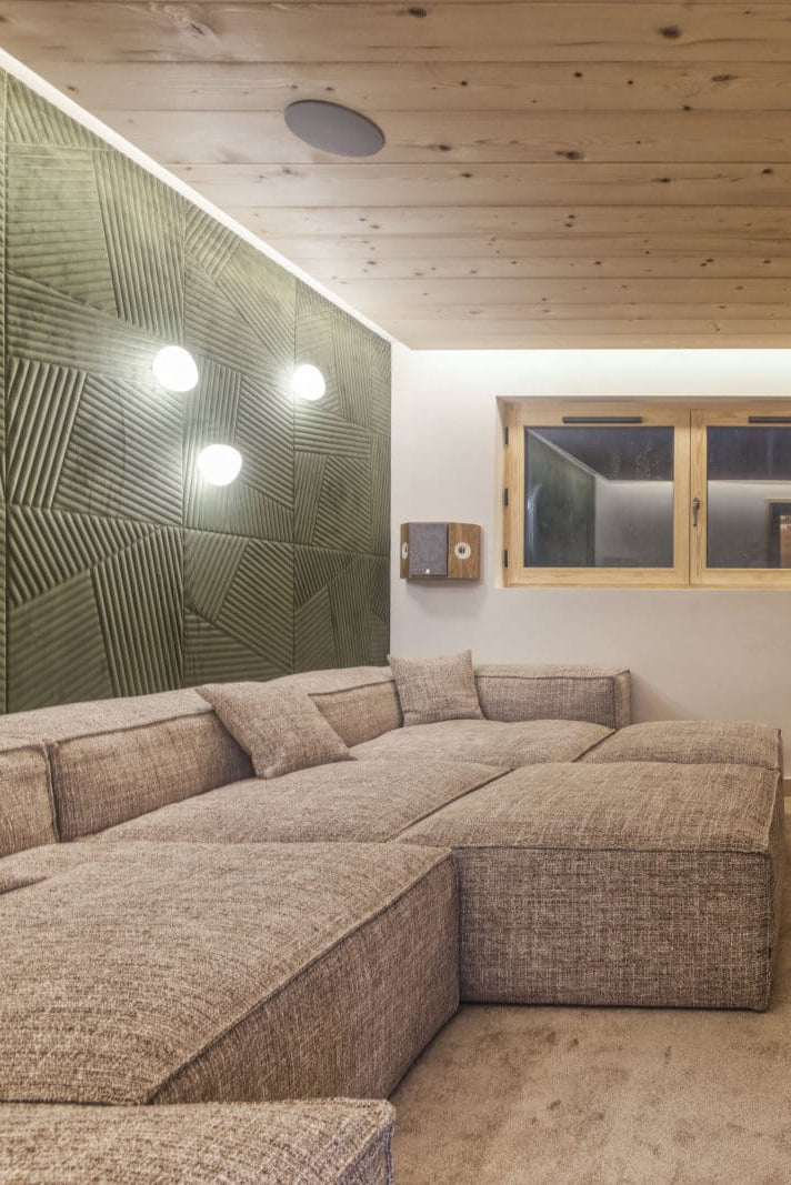 A modern mountain-themed home theater or living room featuring a large sectional sofa, a wooden media console, a wall-mounted TV displaying a mountain landscape, and a textured green accent wall.
