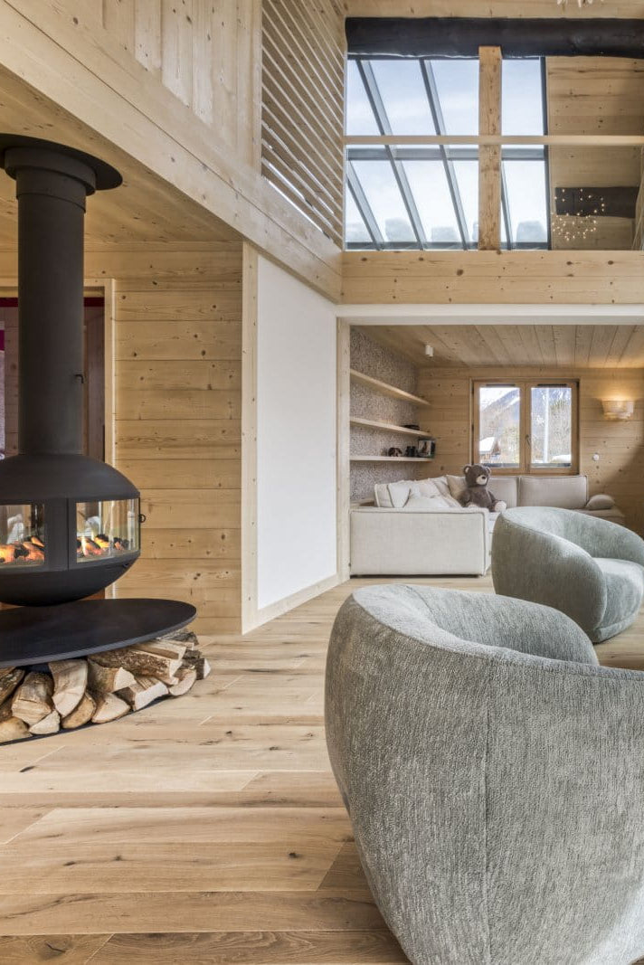 A modern mountain chalet living room with light wood walls and floors, featuring a black freestanding wood-burning stove, two gray upholstered armchairs, a curved sofa, and large windows overlooking snowy mountains.