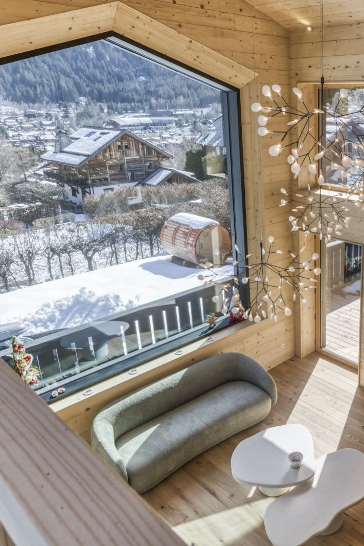 A high-ceilinged wooden chalet interior with a large floor-to-ceiling window overlooking a snowy mountain village. The space features light wood walls and floors, a modern chandelier, a curved gray sofa, a gray armchair, white sculptural side tables, and a dining area in the background.
