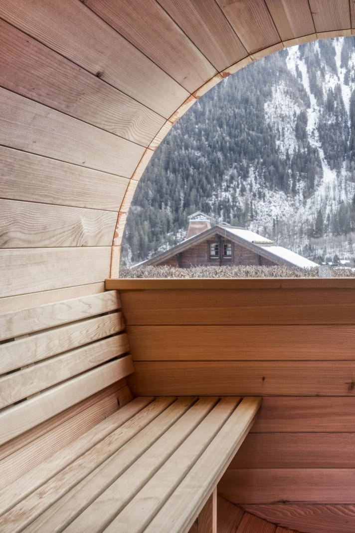 A wooden sauna interior with tiered benches and a large arched window overlooking a snowy mountain landscape.