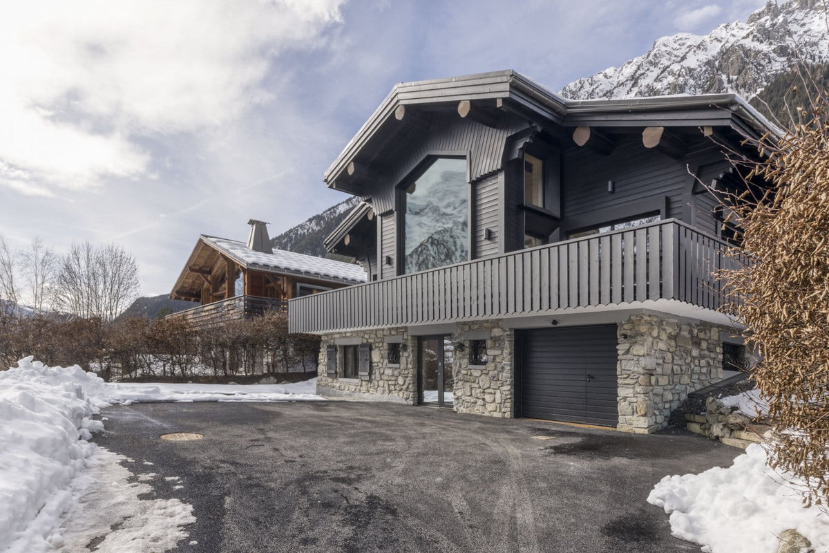 A modern alpine-style chalet mountain house with dark gray wood siding, a stone base, a large wraparound garage door, and snow-covered mountains in the background.