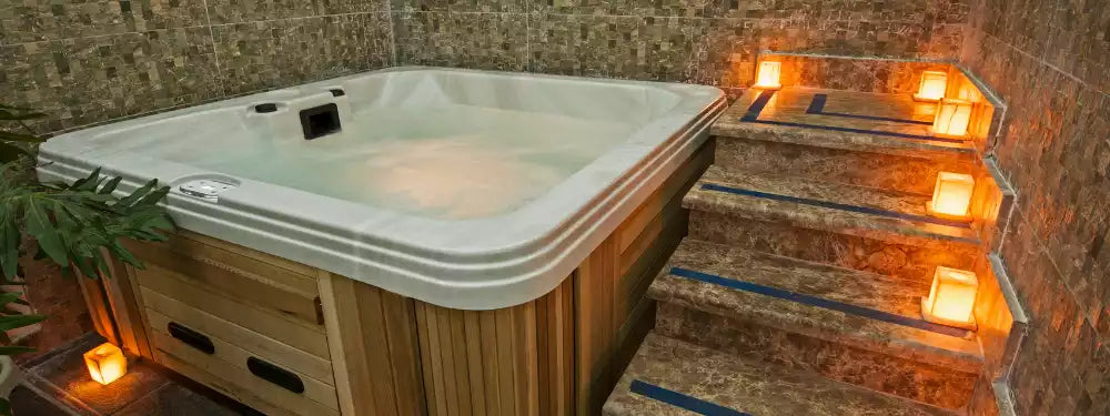 A wooden hot tub with a white shell sits beside stone steps illuminated by warm glowing candles in a tiled indoor setting.