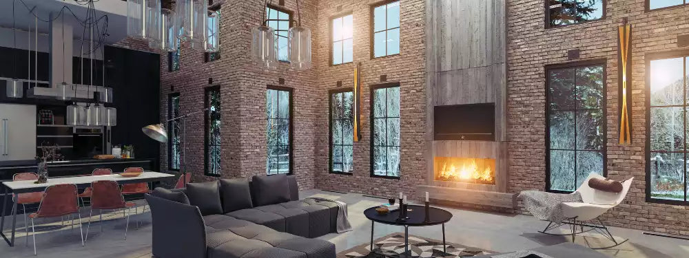 A modern loft-style living room with exposed brick walls, a large gray sectional sofa, a black fireplace with a glowing fire, a dining area with red chairs, and a white lounge chair near tall windows.
