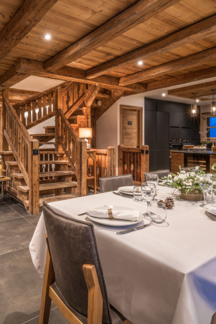 A luxury chalet-style dining room with a wooden staircase, stone fireplace, and a dining table set with white tablecloths and glassware in the foreground.