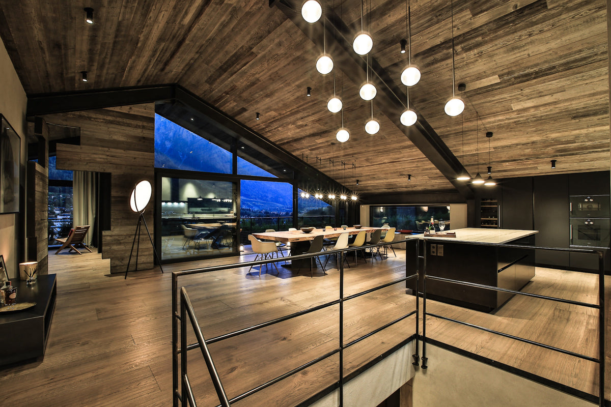 A modern open-plan interior with a high wood ceiling, pendant lights, a dining table, kitchen island, and large windows overlooking a city skyline at dusk.