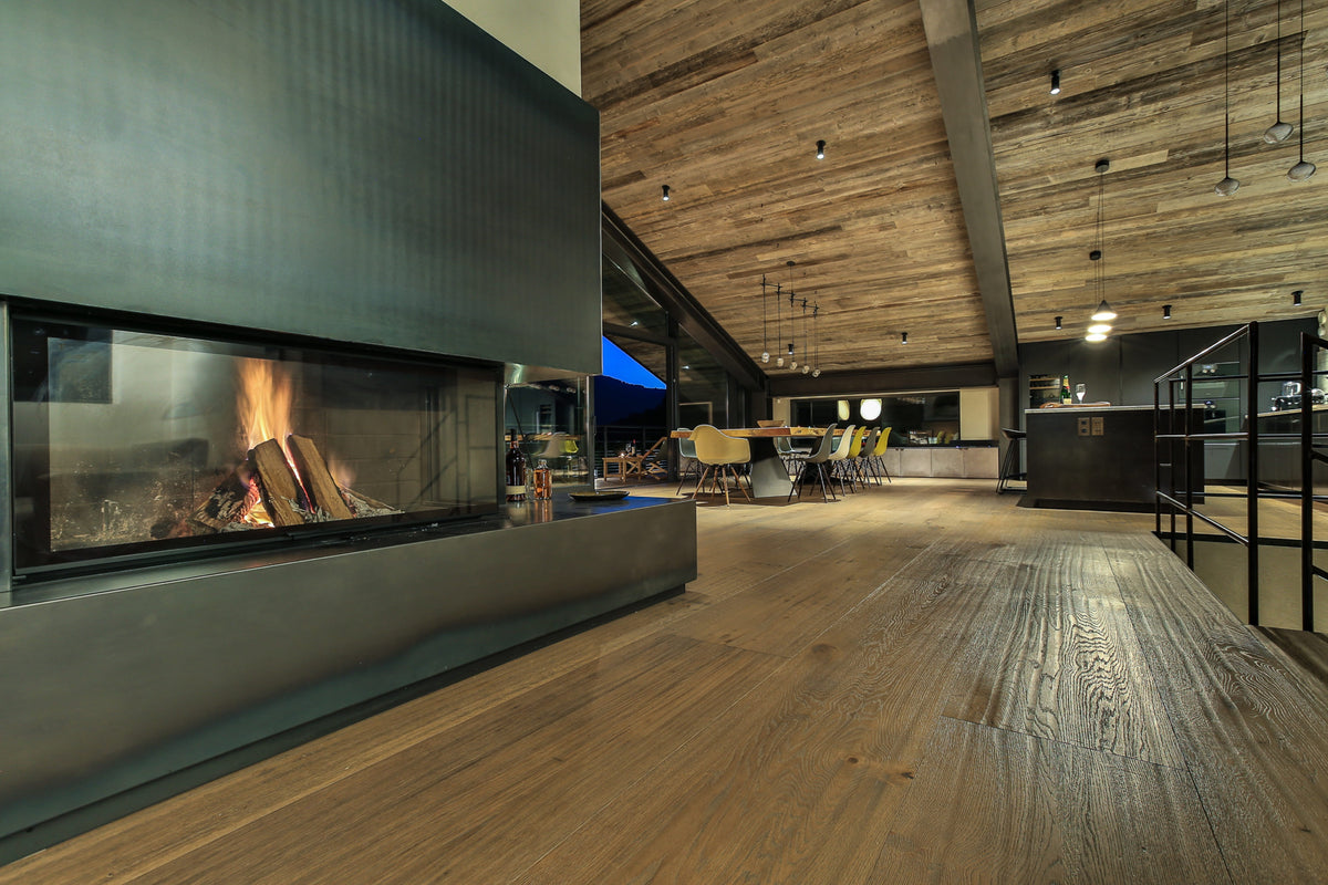A modern wood-burning fireplace with a black metal surround and visible flames, set in a spacious interior with wood flooring, a high wood-paneled ceiling, and an open dining and kitchen area in the background.