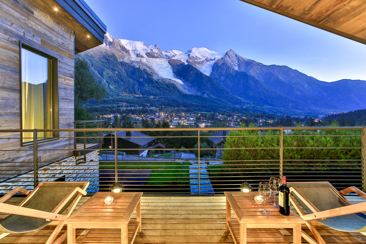 A luxury outdoor balcony overlooking a stunning mountain valley at dusk, furnished with wooden tables, lounge chairs, and a bottle of wine and glasses. The background features majestic snow-capped mountains under a deep blue twilight sky.