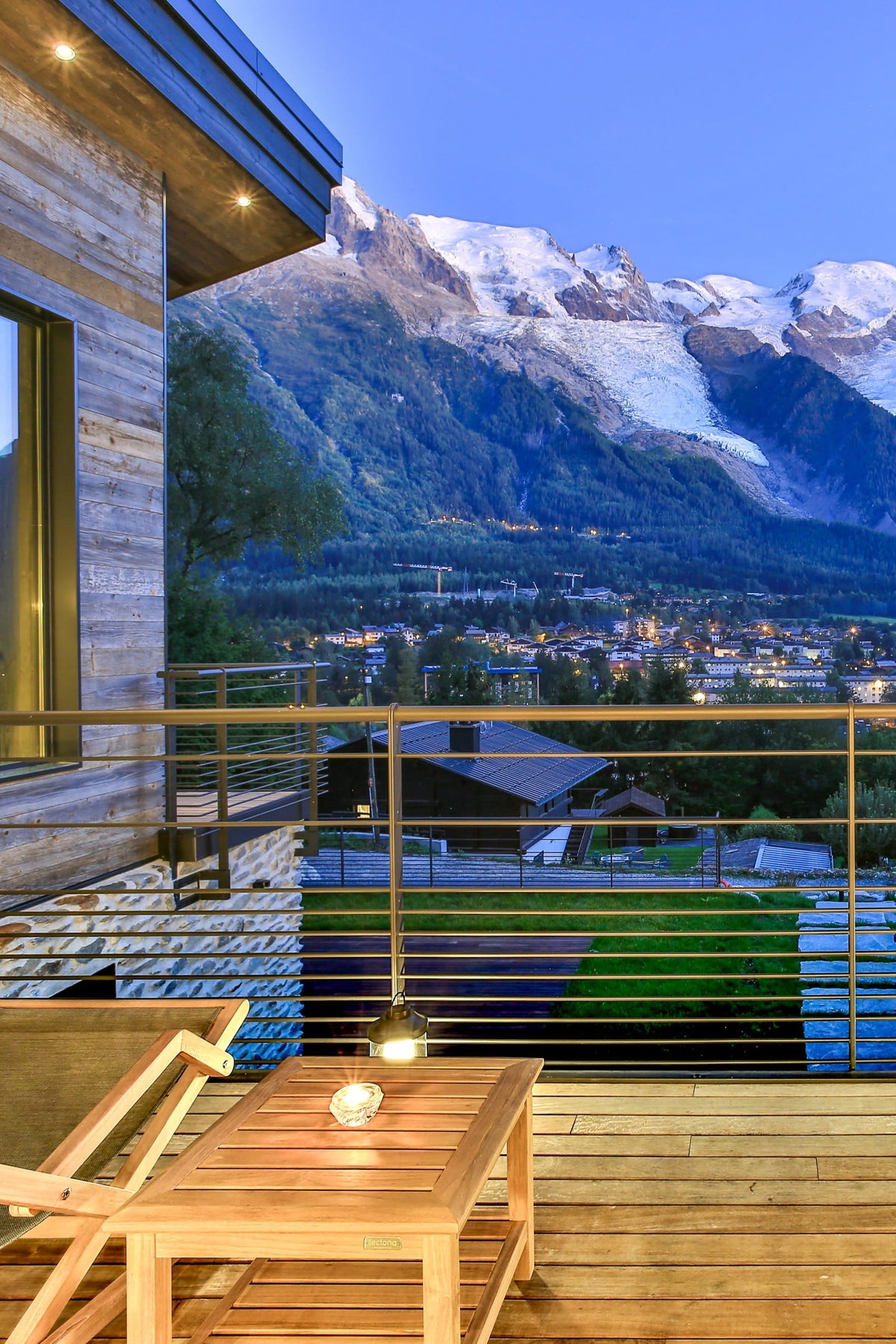 A luxury outdoor balcony overlooking a stunning mountain valley at dusk, furnished with wooden tables, lounge chairs, and a bottle of wine and glasses. The background features majestic snow-capped mountains under a deep blue twilight sky.