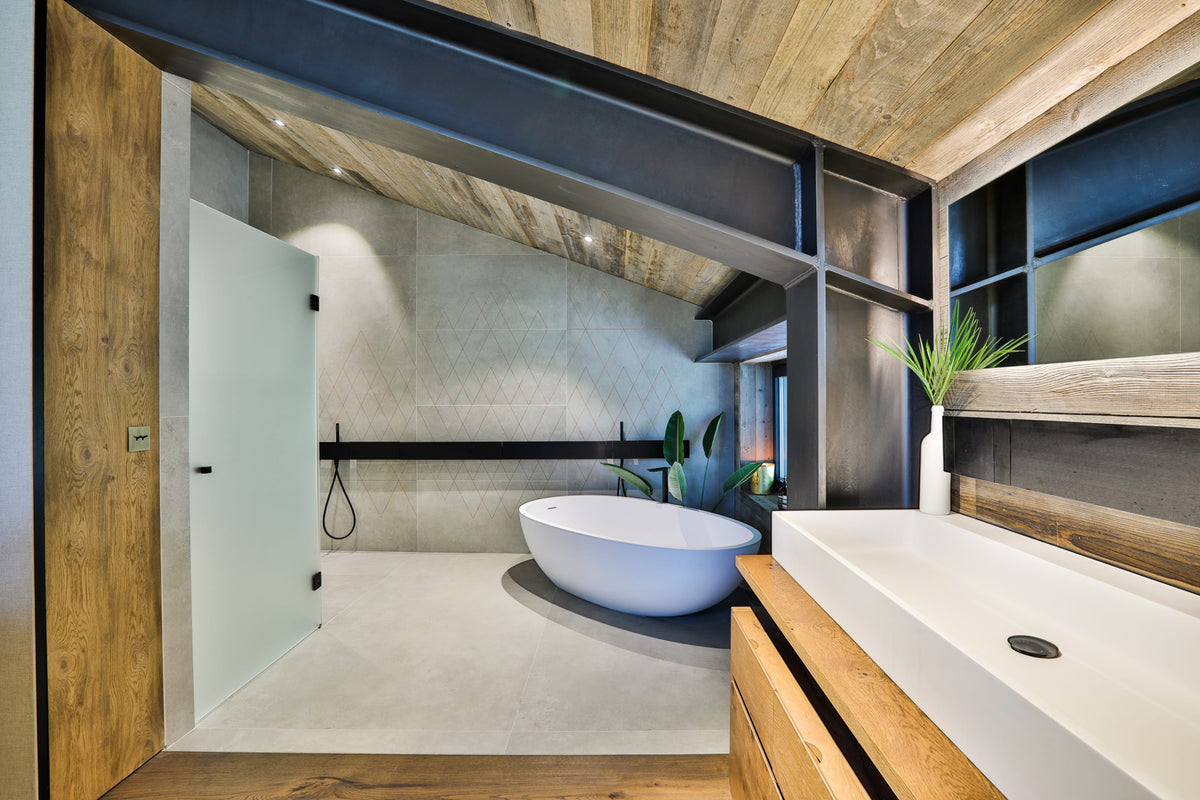 A modern bathroom interior with a freestanding oval bathtub in a tiled shower room, a white sink countertop, wood cabinetry, and a slanted wood ceiling.
