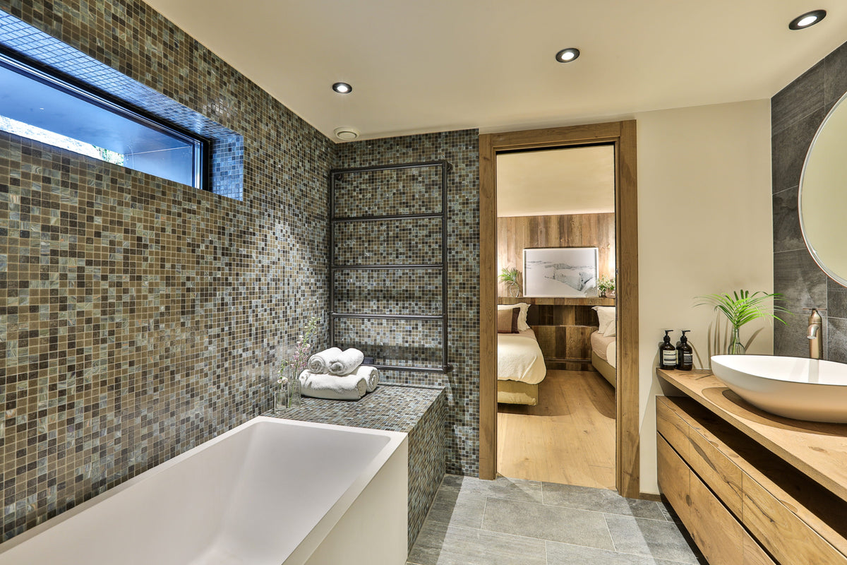 A modern bathroom interior featuring a white freestanding bathtub set against a brown and gray mosaic tile wall, with a wooden vanity and vessel sink on the right. An open wooden door reveals a bedroom in the background.