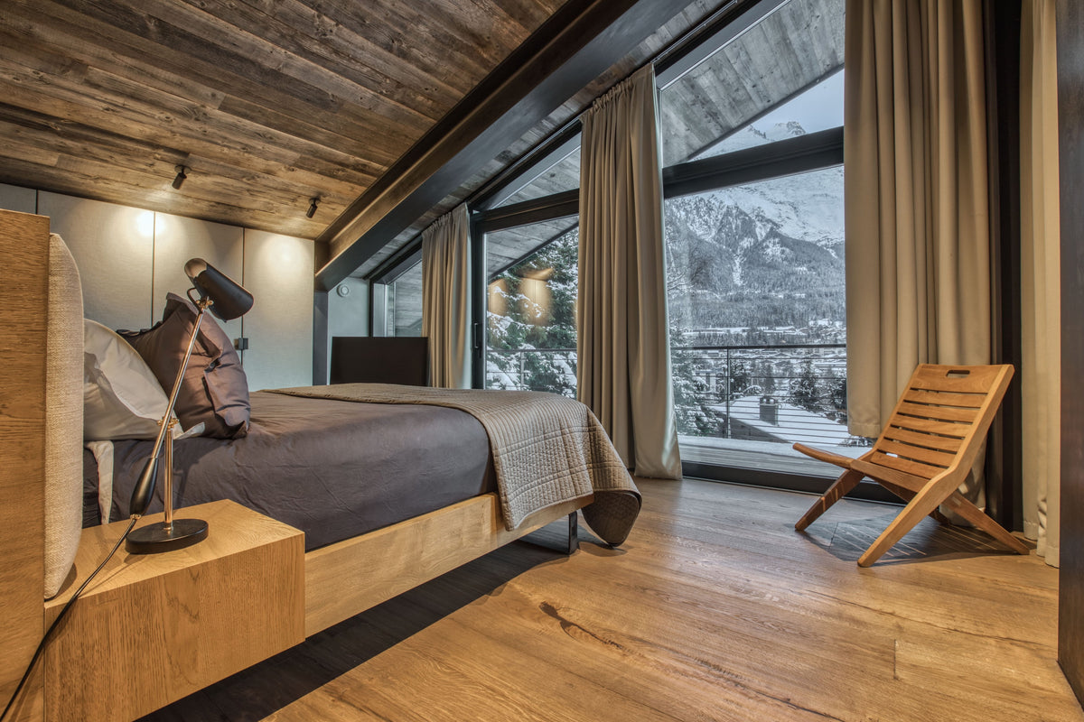 A modern mountain-view bedroom with a low wooden bed, gray bedding, a bedside lamp, a wooden chair by large windows, and a rustic wood ceiling.