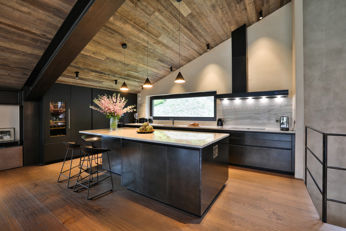 Modern kitchen interior with a large central island, dark cabinetry, wood flooring, exposed wood ceiling beams, pendant lights, and a stone backsplash. No people or animals are present.