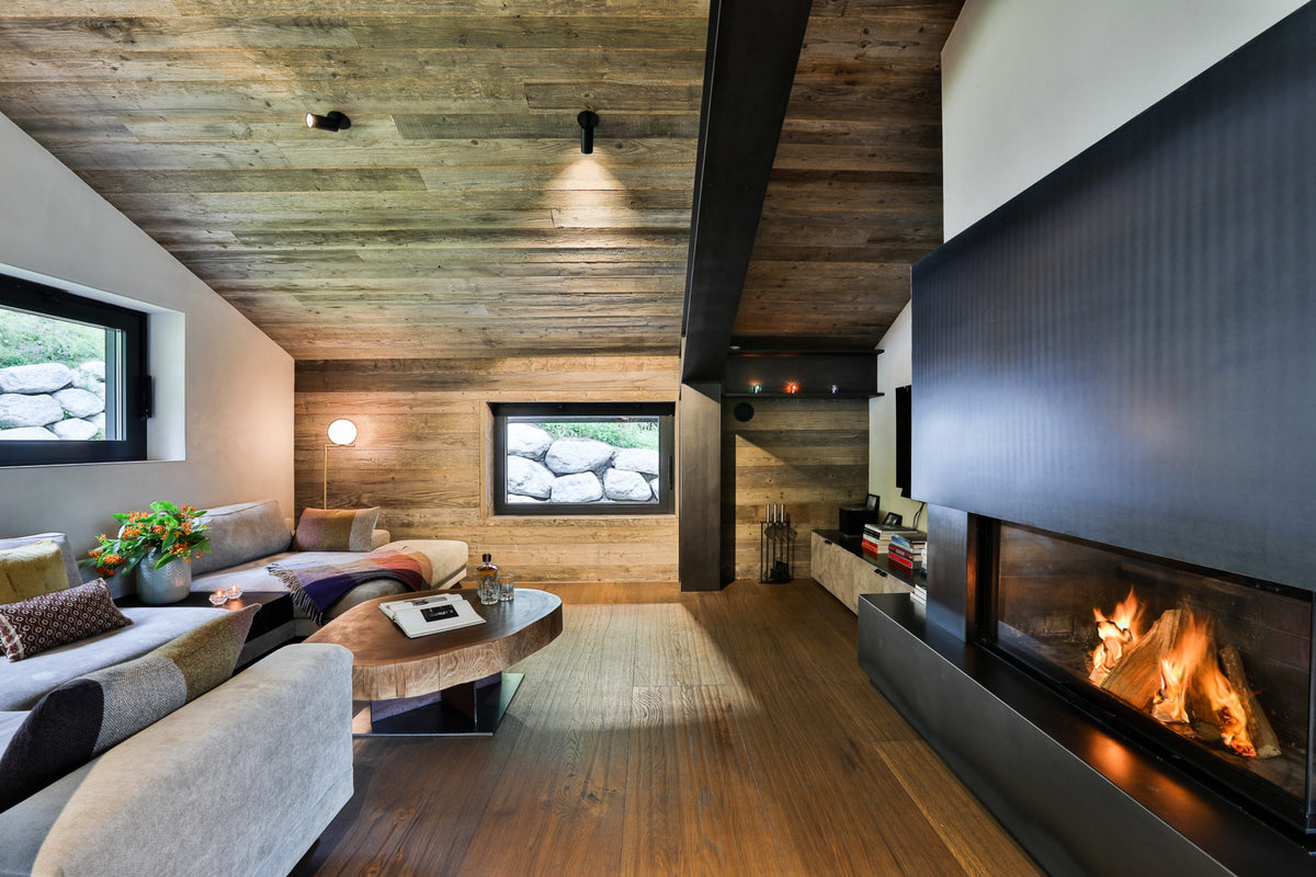 A modern living room interior with a large black fireplace wall and visible fire, wood-paneled ceiling and walls, a gray sofa, and a round wooden coffee table.