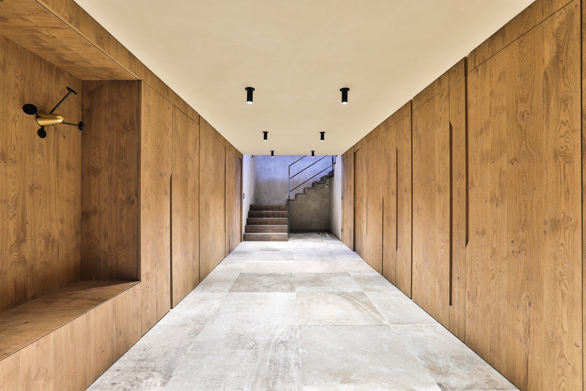 A long interior hallway with light wood paneling on both sides, a pale stone floor, recessed ceiling lights, and a staircase visible at the far end.