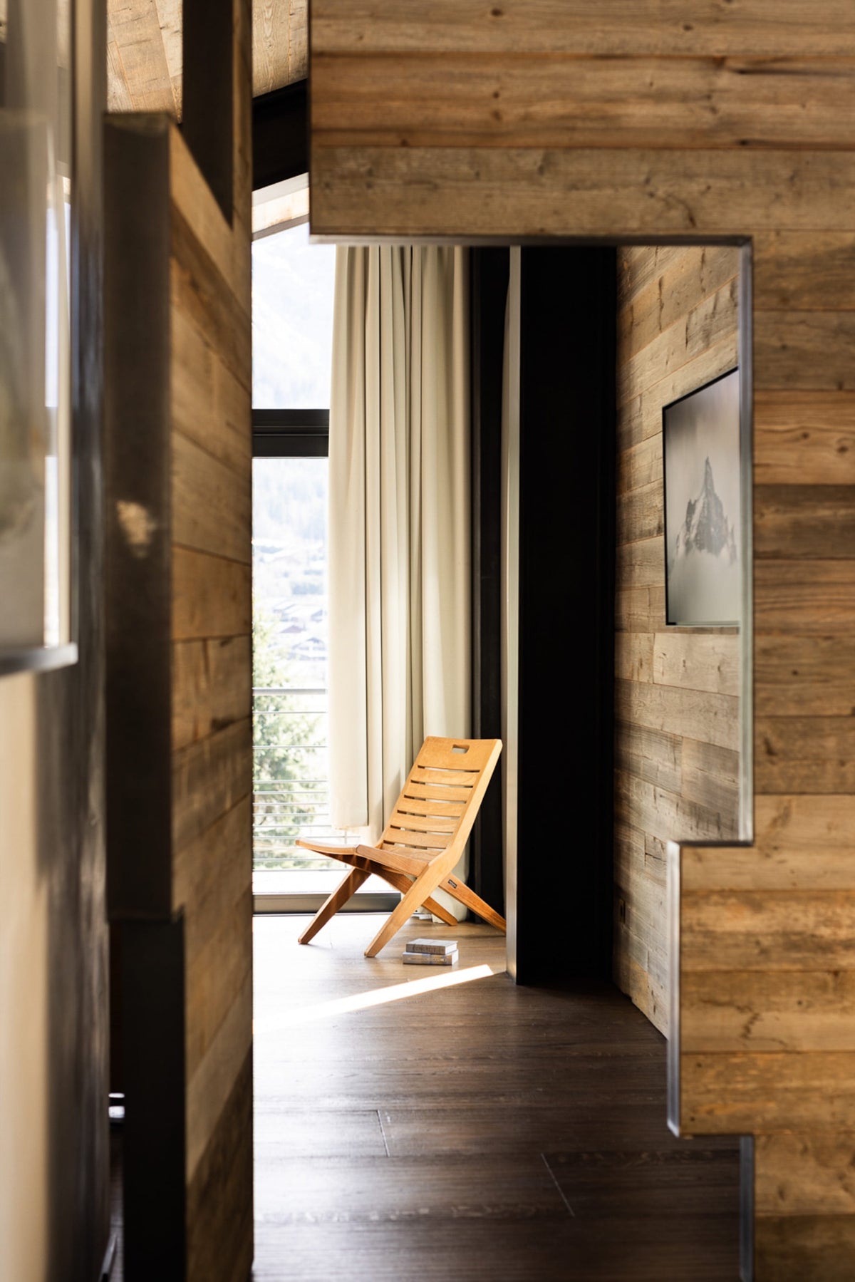 A modern interior hallway with wood-paneled walls and floor, leading to a sunlit room with a wooden folding chair and a large window with cream curtains.