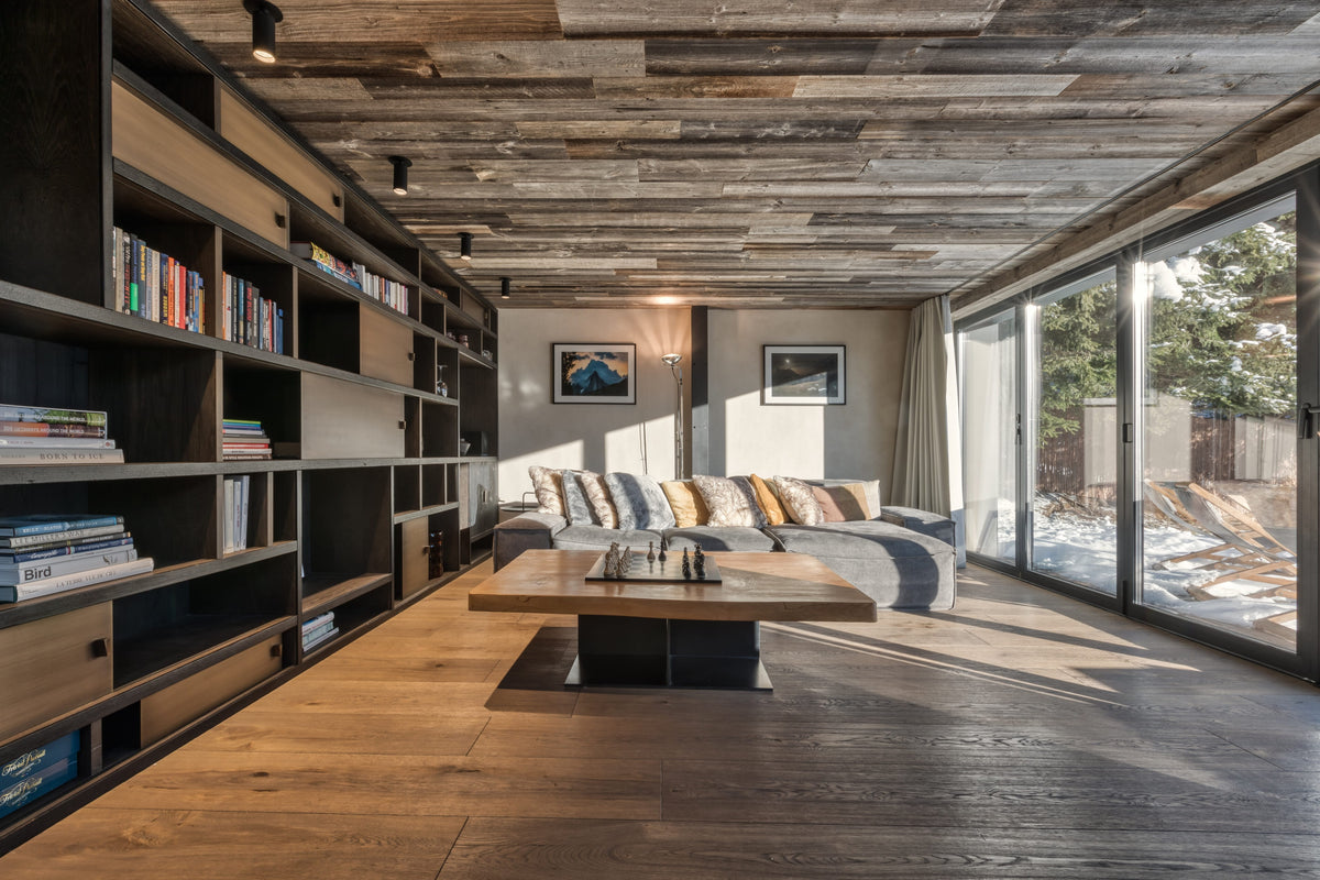 A modern rustic living room with a rustic wood plank ceiling, dark wood bookshelves on the left, a low wooden coffee table in the center, and a gray sofa with colorful striped pillows. Large floor-to-ceiling windows on the right show a snowy outdoor landscape.