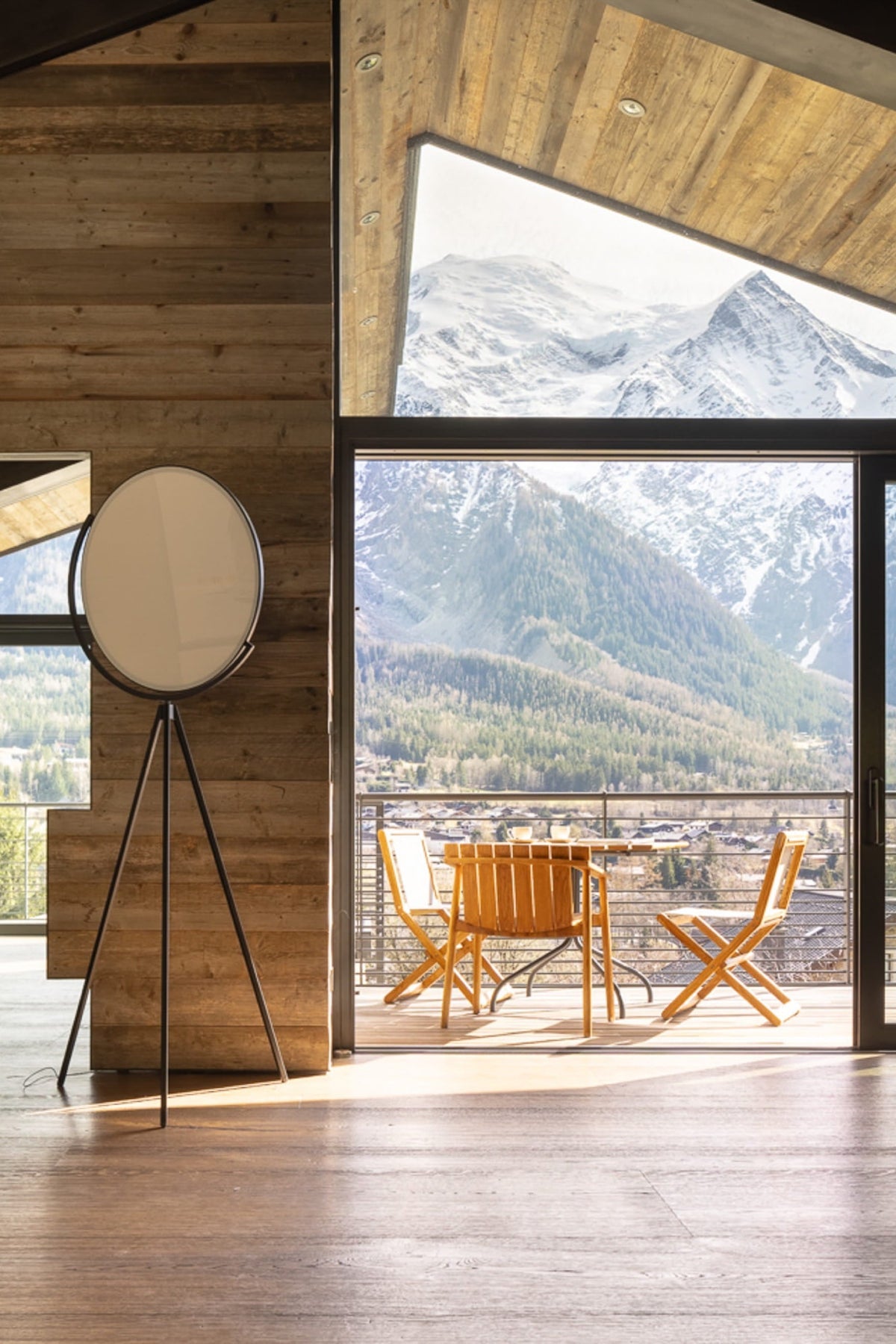 A modern interior room with exposed wood walls and large floor-to-ceiling windows overlooking snow-capped mountains. A round floor mirror on a tripod stand, wooden dining chairs, and a dining table are visible in the room.