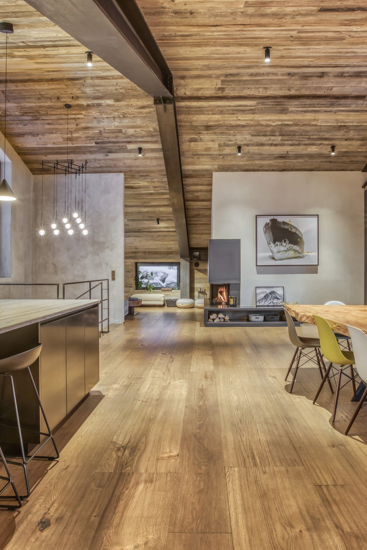 A modern open-plan interior with a large wood dining table and chairs, a stainless steel kitchen island with bar stools, exposed wood ceiling beams, and a fireplace in the background.
