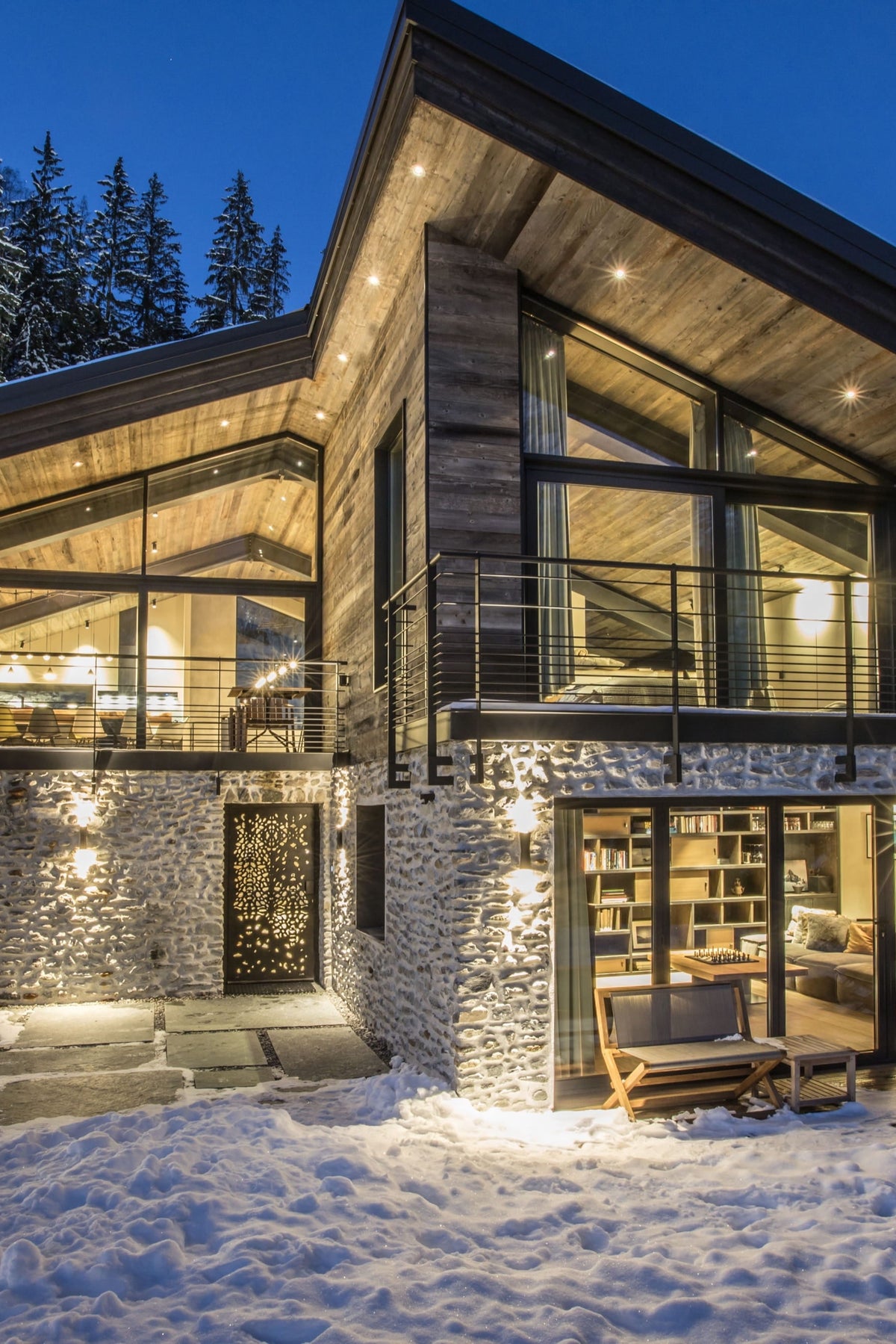 Modern luxury chalet-style mountain house with stone and wood exterior, large floor-to-ceiling glass windows, snow-covered ground, and evergreen trees in the background at dusk.