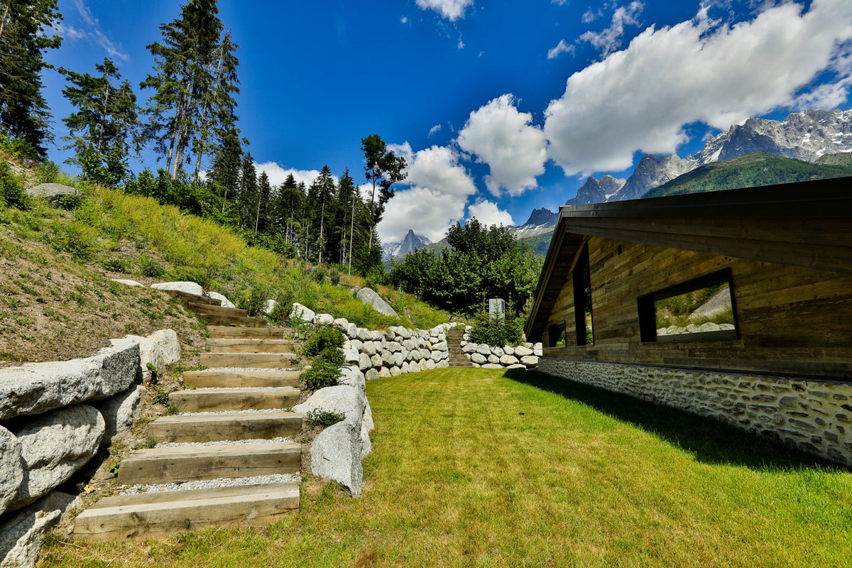 A scenic mountain landscape with a wooden building, stone steps, grassy slopes, trees, and a bright blue sky with white clouds.