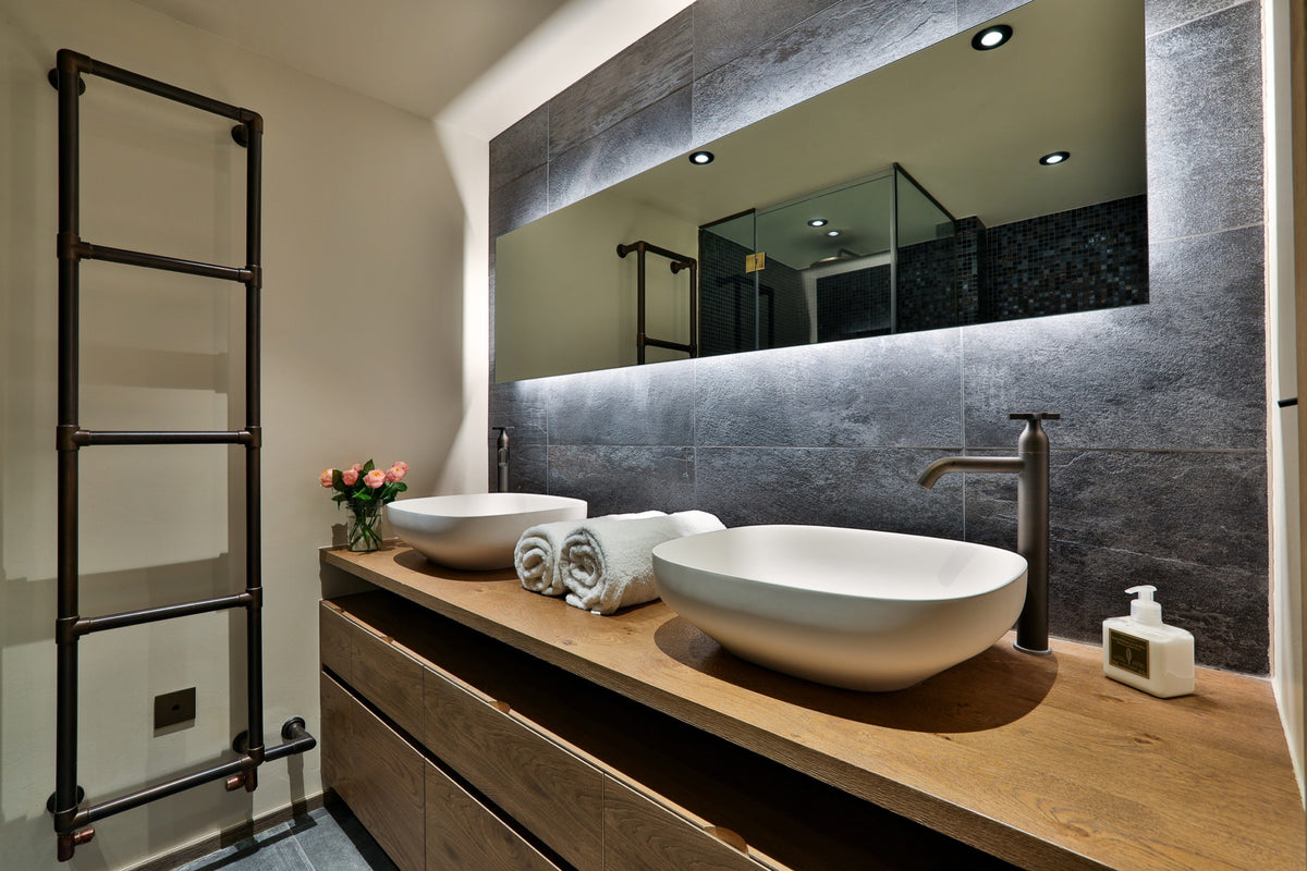 Modern double bathroom vanity with two white vessel sinks on a wooden countertop, a large backlit mirror, dark gray stone wall paneling, and a black metal towel ladder.