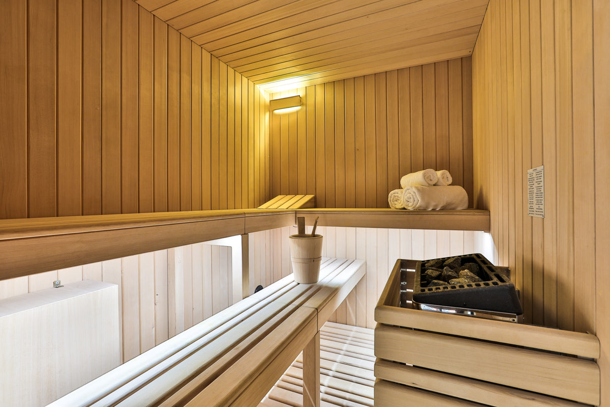 A warm wooden sauna interior with tiered wooden benches, a hanging towel, a small plant in a pot, and a black sauna stove on the right side.