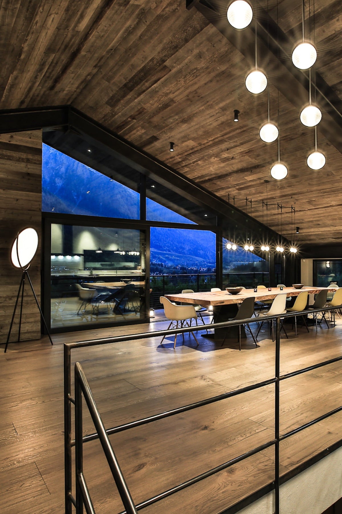 A modern open-plan interior with a high wood ceiling, pendant lights, a dining table, kitchen island, and large windows overlooking a city skyline at dusk.