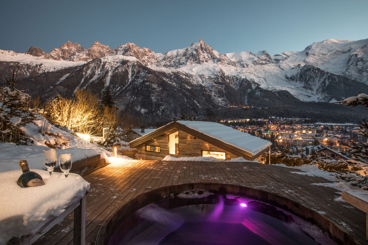 A luxury mountain chalet with a hot spring outdoor pool in the foreground, set against a backdrop of snow-covered alpine peaks and a glowing valley town at dusk.
