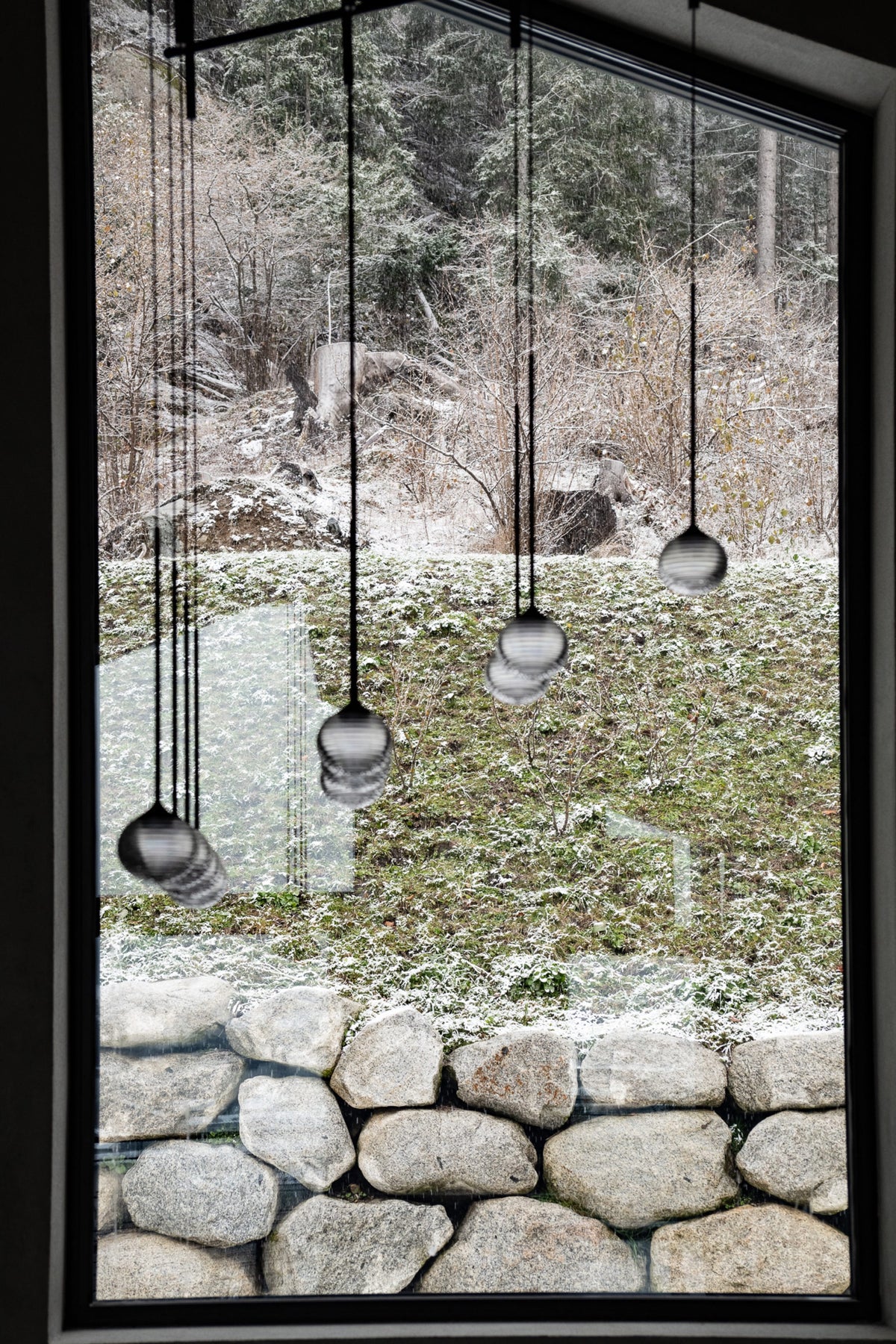 A large black-framed window overlooking a snowy outdoor landscape with trees, a stone wall, and hanging spherical pendant lights in the foreground.