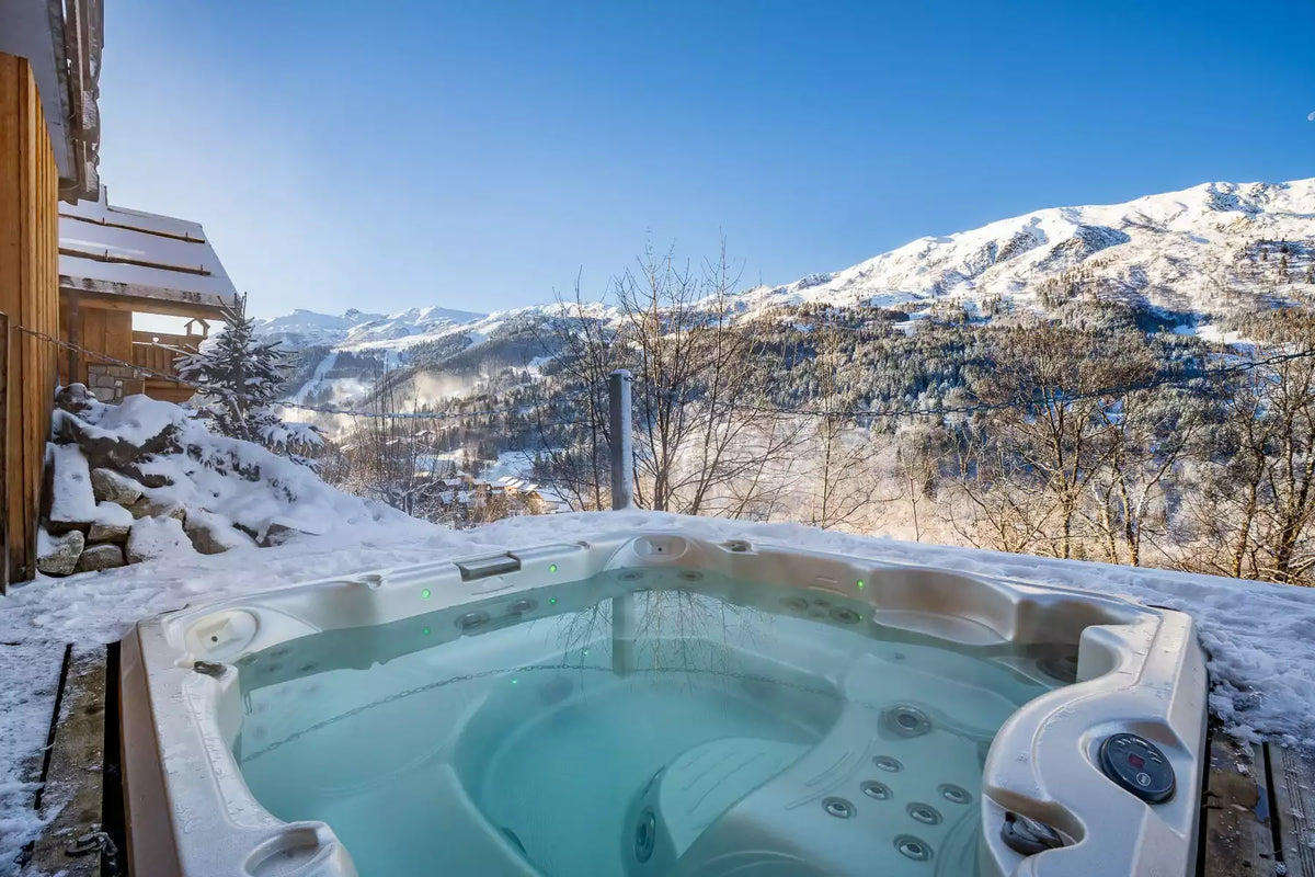 An outdoor hot tub filled with clear blue water, situated on a snowy mountain terrace with a dramatic snow-covered mountain range and blue sky in the background.
