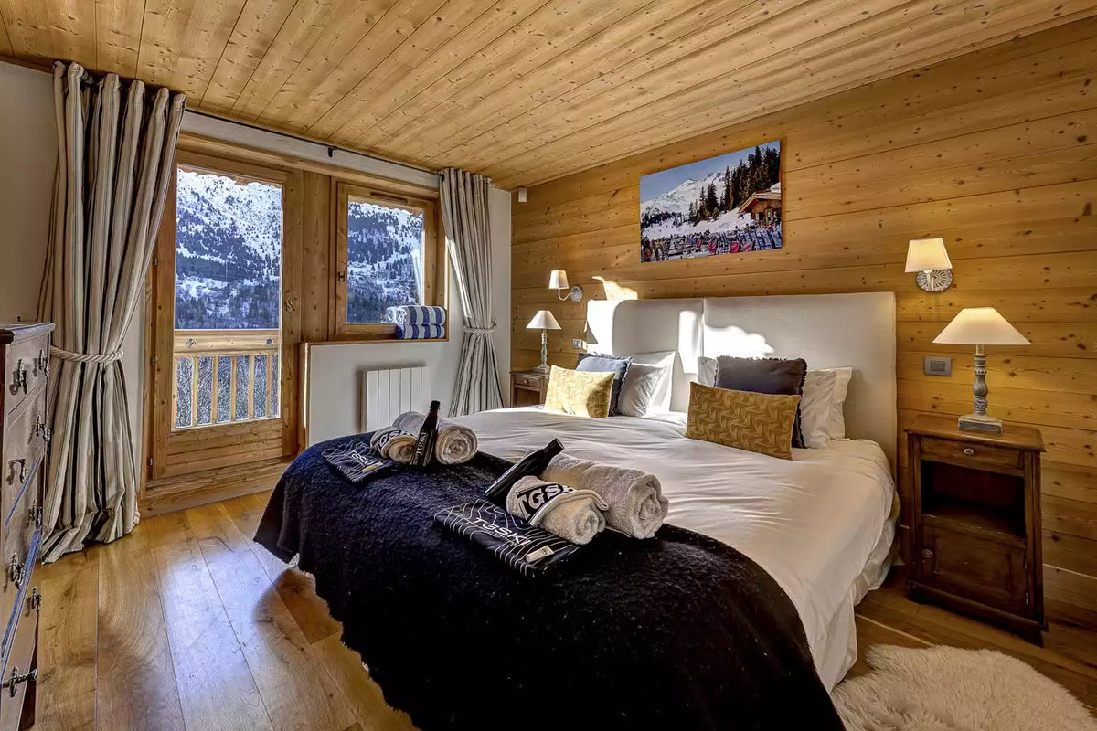 A cozy alpine-style bedroom with a large bed dressed in white linens and a dark throw blanket, set against warm wood-paneled walls and ceiling. Two windows with gray curtains look out to a snowy mountain scene, and wall-mounted lamps and a framed landscape print enhance the rustic lodge ambiance.