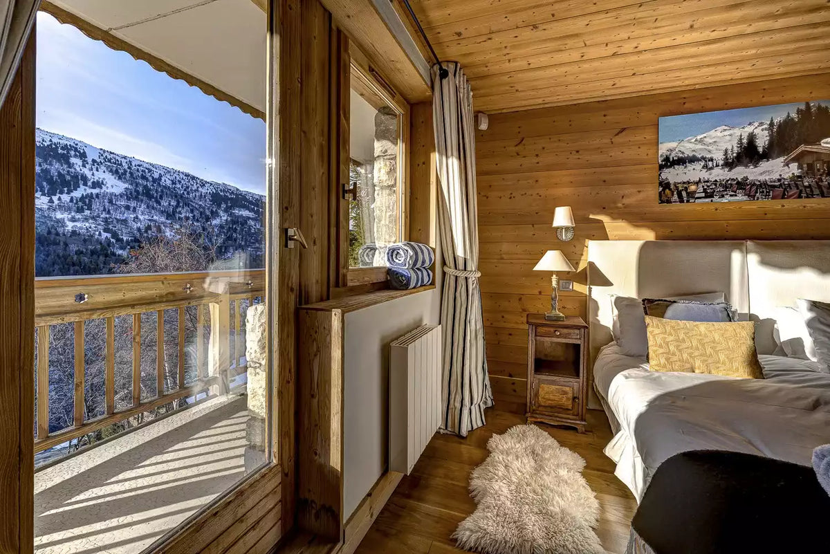 A cozy wooden chalet bedroom with a large window overlooking a snowy mountain landscape, featuring a bed with gray bedding, a wooden nightstand, and a framed mountain print on the wood-paneled wall.