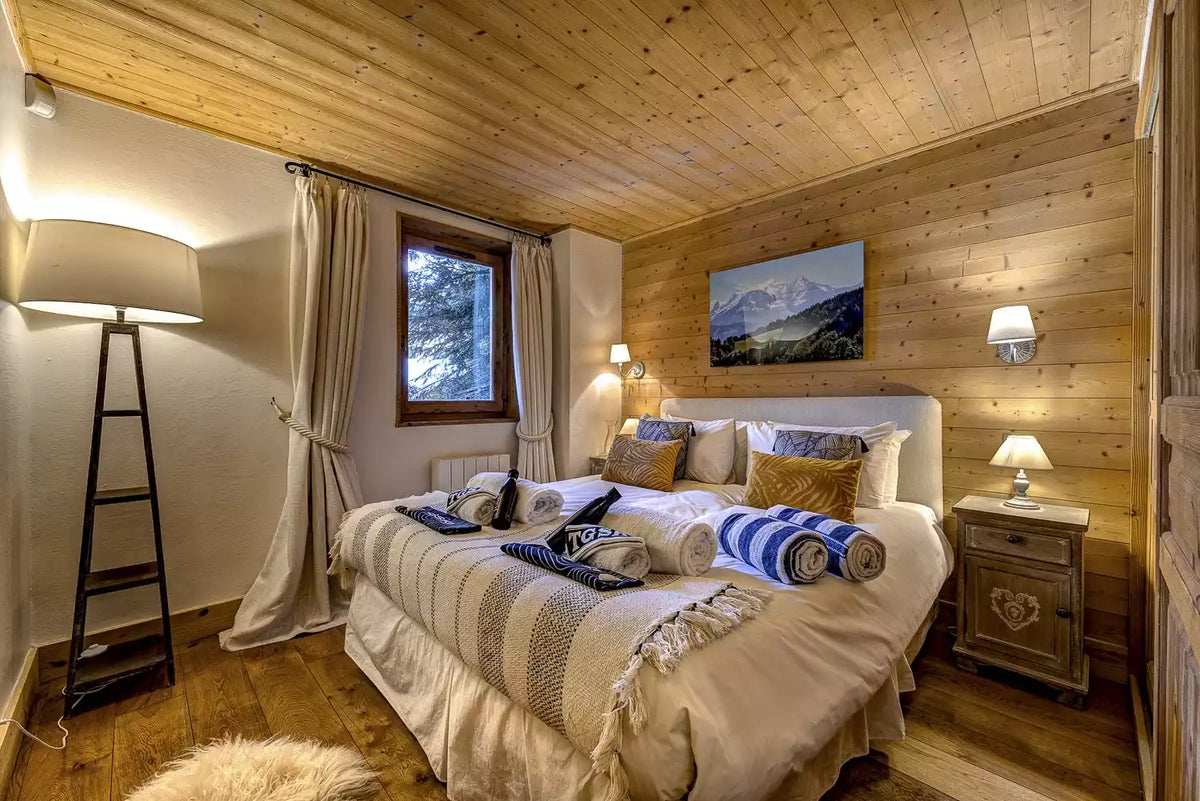A cozy mountain-style bedroom with wood-paneled walls and ceiling, featuring a neatly made bed with cream bedding, blue striped pillows and rolled towels, a window with beige curtains, bedside tables with lamps, and a framed landscape painting.