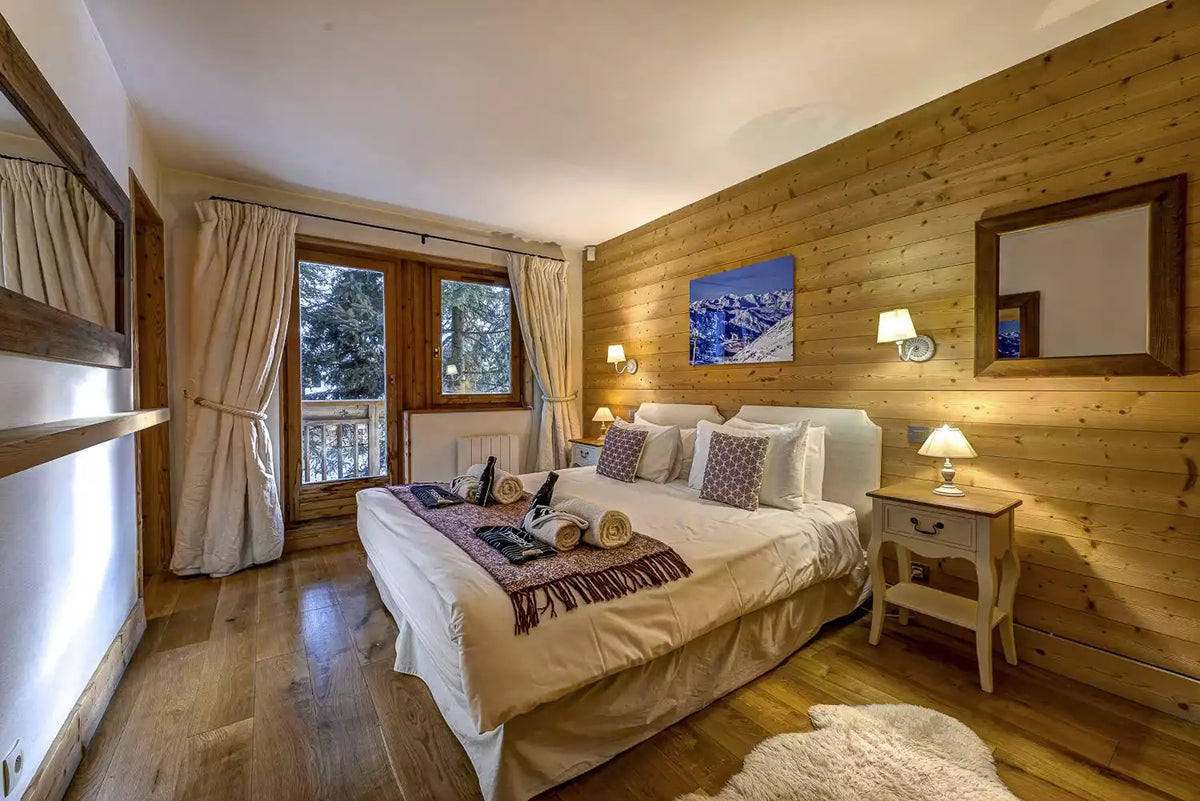 A cozy, rustic mountain-style bedroom with a large bed dressed in cream bedding and purple patterned pillows, set against a warm wood-paneled wall. The room features a matching wooden nightstand, wall sconces, a mirror, and a window with curtains looking out to trees.