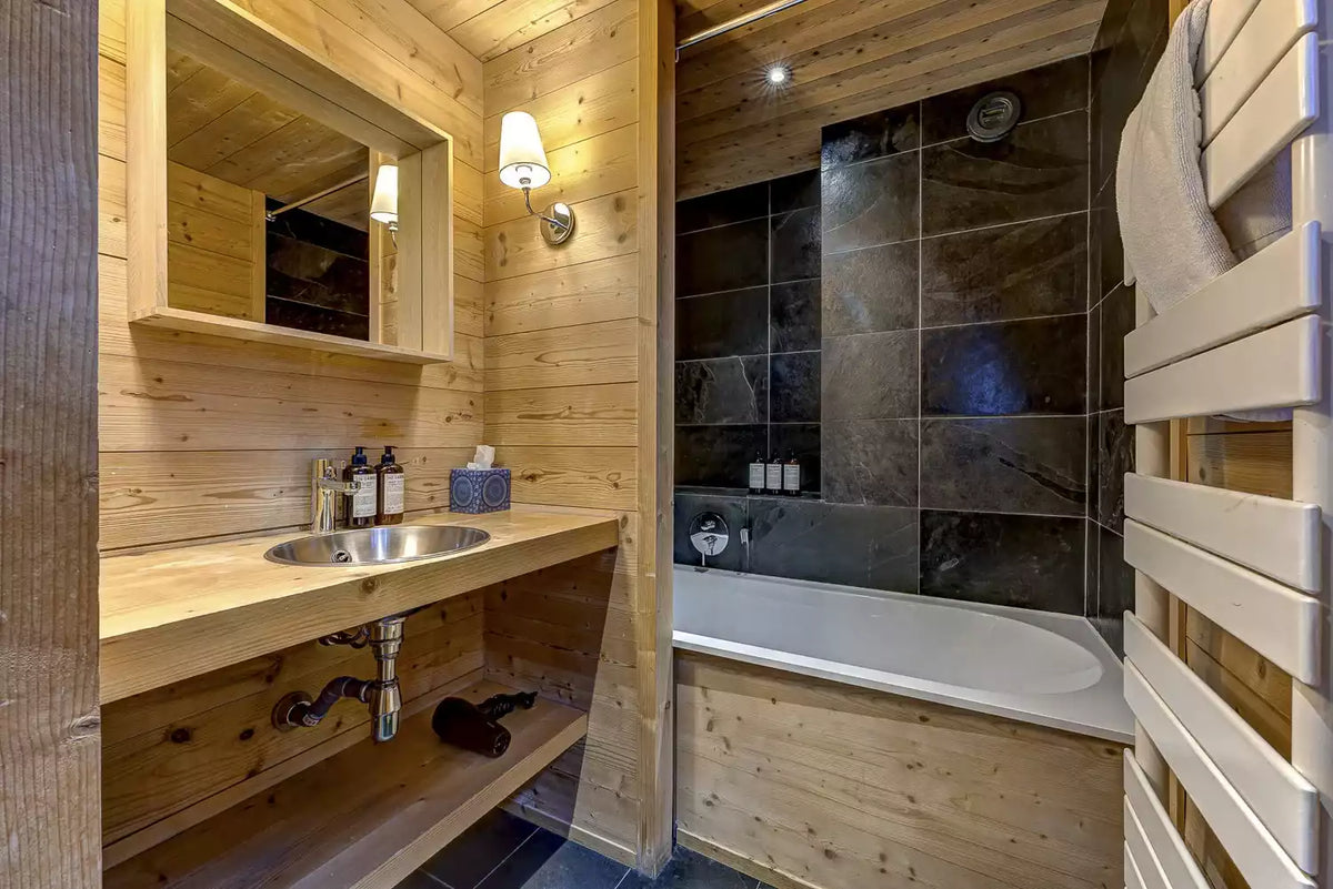 A rustic modern bathroom with light wood paneling, a wooden vanity with a sink, a white bathtub with dark tiled walls, and a wooden towel rack with a white towel.