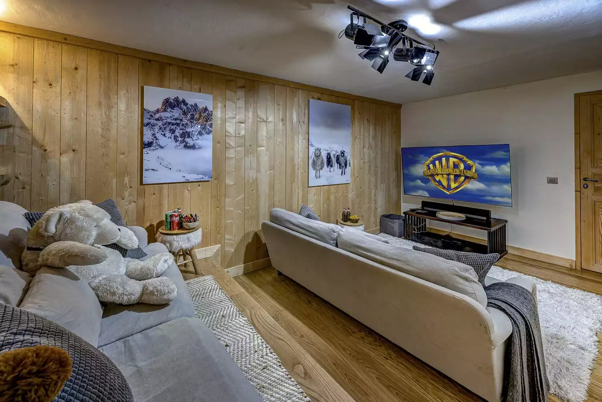 A modern home theater setup in a wood-paneled room, featuring a light gray sectional sofa, plush white stuffed animal, and a large flat-screen TV mounted on the back wall displaying the Warner Bros. logo.