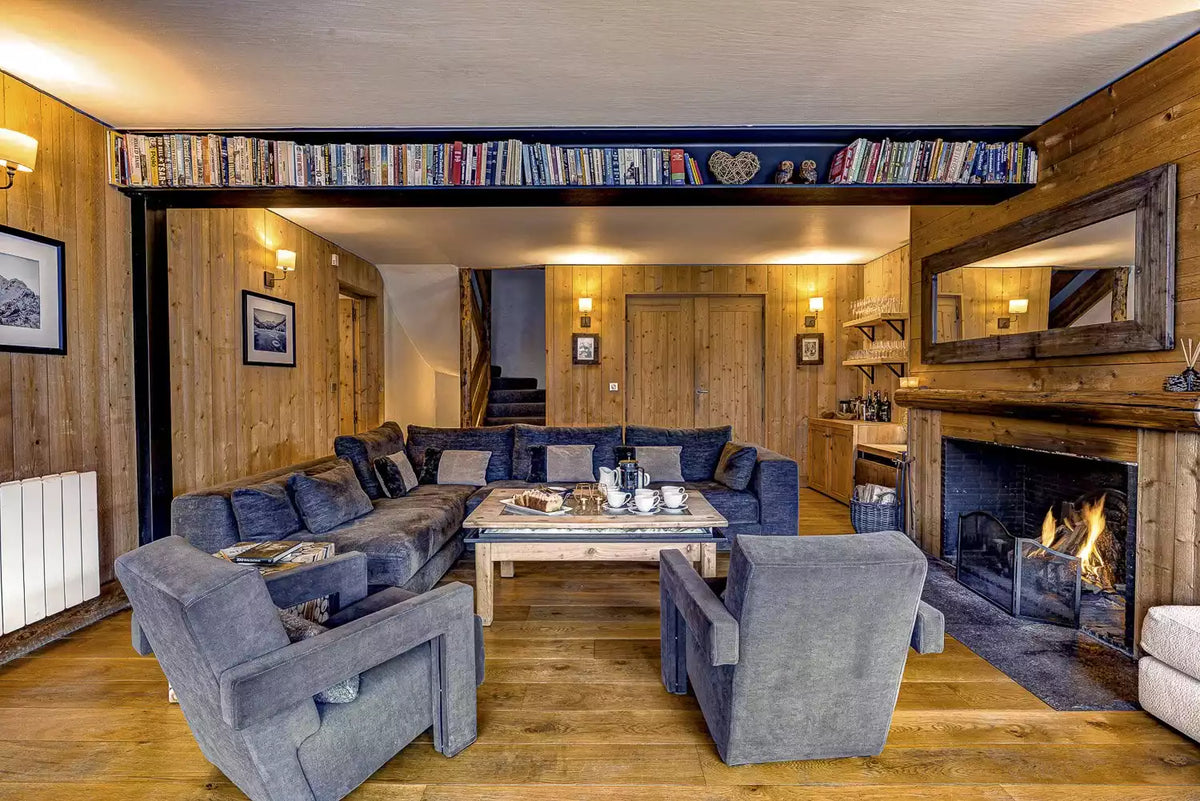 A cozy, rustic living room with wood-paneled walls, a large blue sectional sofa, two matching gray armchairs, a wooden coffee table, and an active fireplace with a large mirror above it.