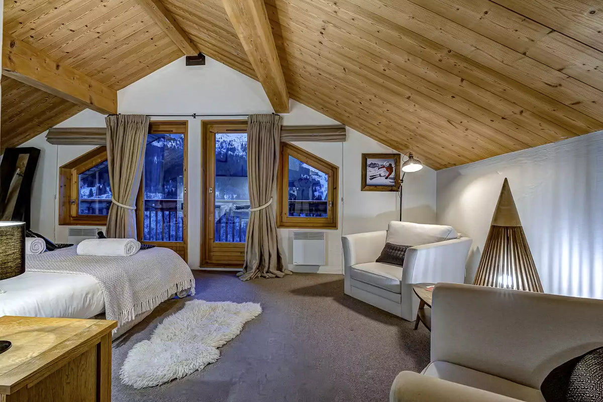 A cozy ski chalet bedroom with a wooden slatted bed, white bedding, a white armchair, a wooden floor lamp, and a slanted wooden ceiling.