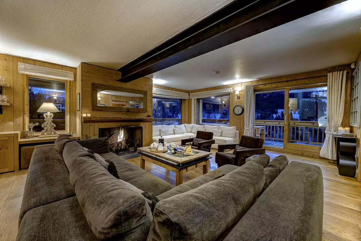 A cozy, rustic mountain chalet living room featuring a large dark brown sectional sofa in the foreground, a wooden fireplace with a mirror, a wooden coffee table, and white sofas in the background near large windows looking out onto a balcony at dusk.