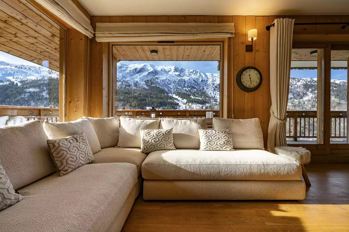 A large U-shaped sectional sofa in cream bouclé fabric with patterned accent pillows, set inside a rustic wooden chalet with large windows overlooking a snowy mountain landscape.