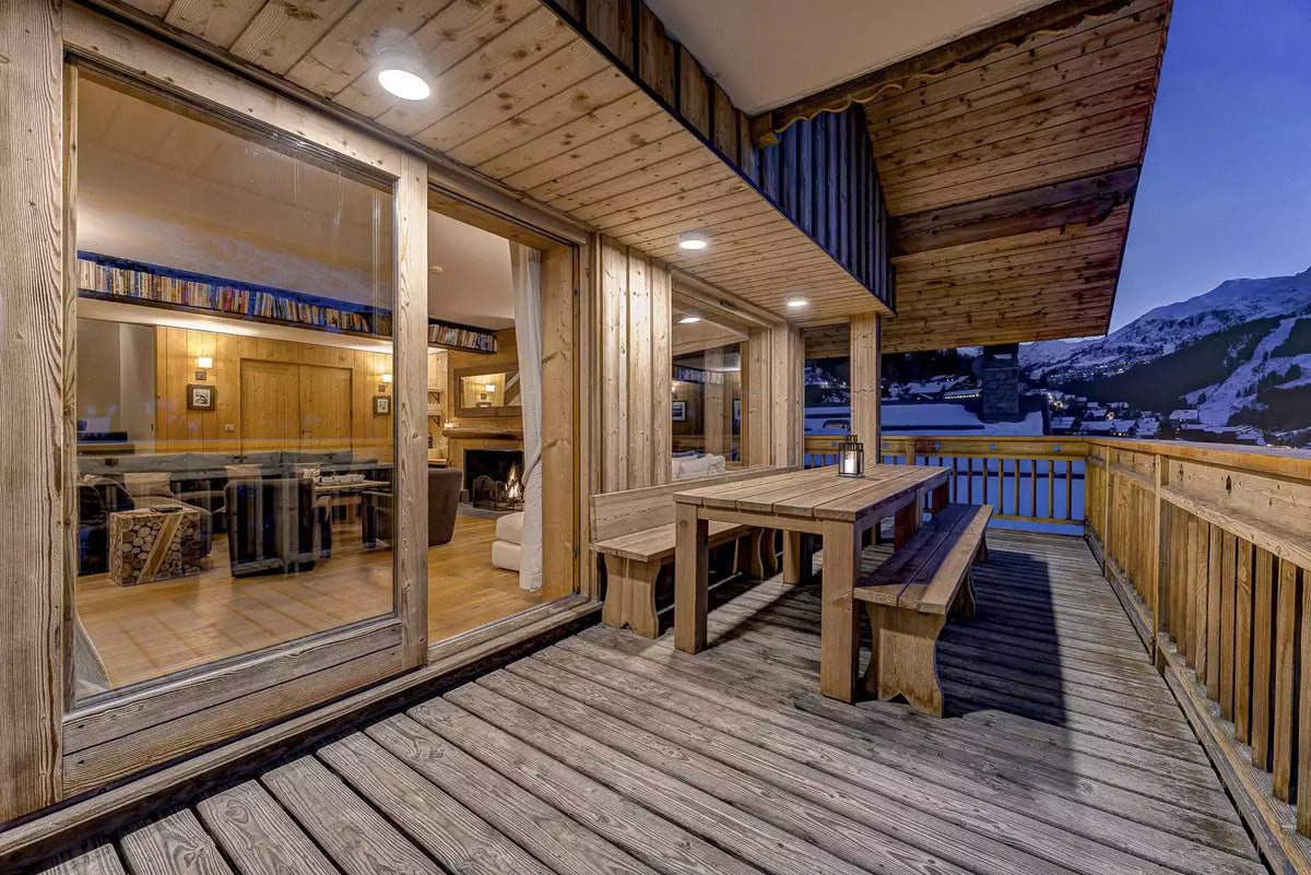 A wooden chalet balcony with a dining table and benches, large glass doors opening to a warm kitchen interior, and a snowy mountain landscape visible at dusk.